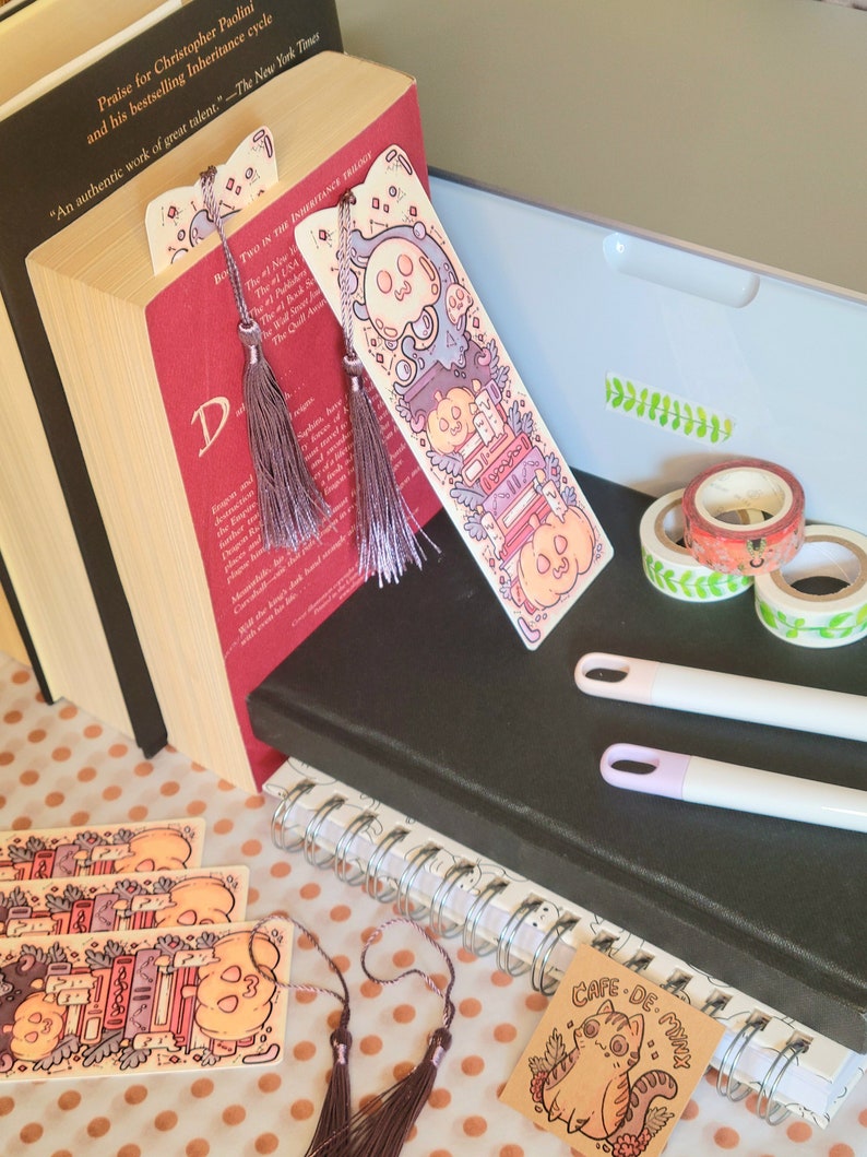 Cute cardstock paper bookmark with tassel and cat ear cut on the top leaning against a red book. The bookmark has an illustration of stacked books, pumpkins, ghosts, and cauldron surrounded by plants. It is surrounded by other books, pens, etc