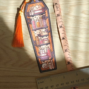 Gold orange coffin shaped bookcase bookmark with orange tassel. Features books, potions, pumpkin, candles and other witch library designs on the shelves.  It rests on a light wood table with ruler beside to show size (roughly 6inches by 2inches).