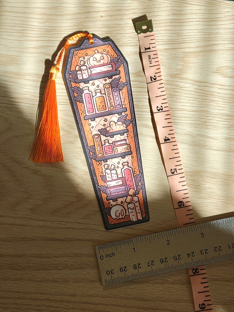 Gold orange coffin shaped bookcase bookmark with orange tassel. Features books, potions, pumpkin, candles and other witch library designs on the shelves.  It rests on a light wood table with ruler beside to show size (roughly 6inches by 2inches).