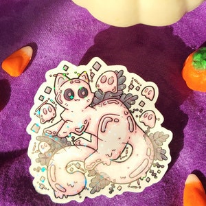 Holographic halloween ghost cat sticker. It is a light blue, white ghost surrounded by plant motifs and floating ghost friends. It is sitting on a purple cloth with pumpkin and candy corn decorations surrounding it.