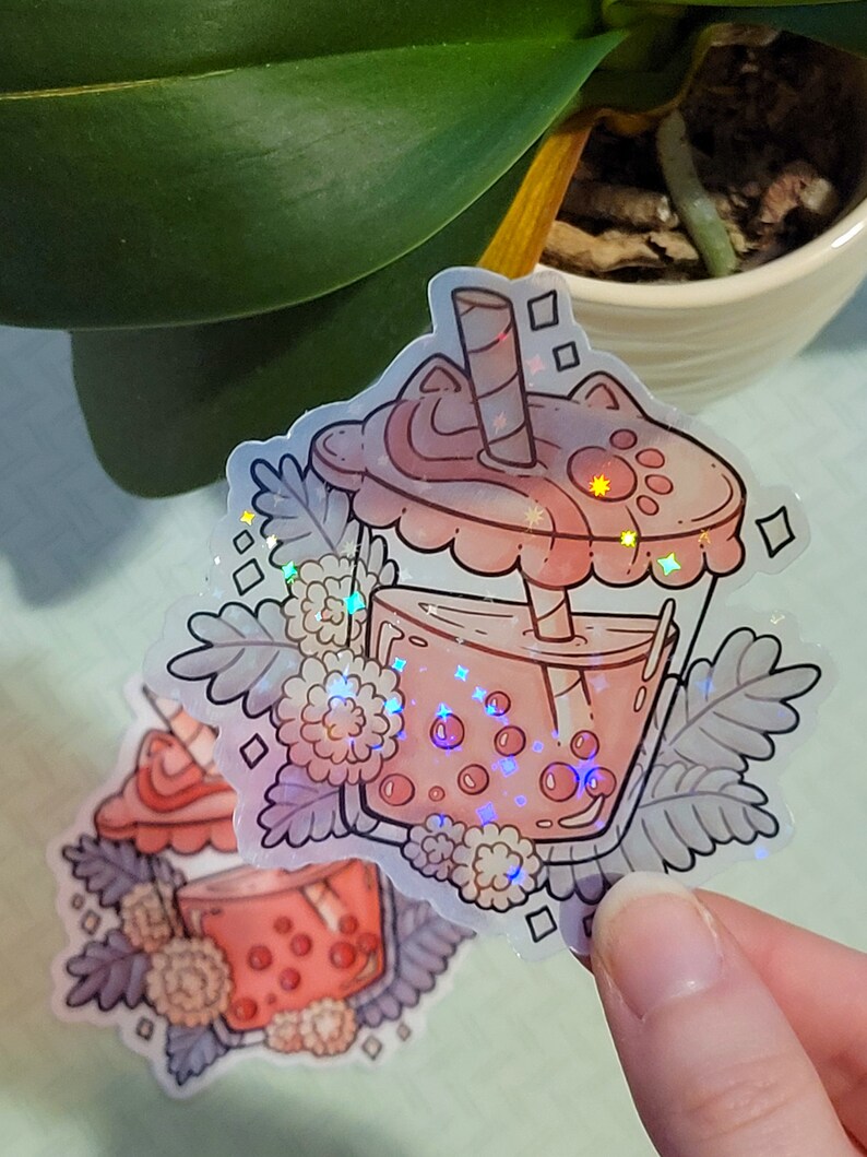 K&ouml;nnte beinhalten: Ein holografischer Aufkleber mit einem rosa Bubble Tea mit einem Katzenkopfdeckel, einem Strohhalm und Boba-Perlen. Der Aufkleber ist von wei&szlig;en Blumen und Bl&auml;ttern umgeben.