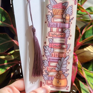 Two cute cardstock paper bookmarks with tassel and cat ear cut on the top held in a hand with a plant behind. One bookmark has an illustration of aesthetic stacked pink and purple books surrounded by pretty plants and the other the white backside.