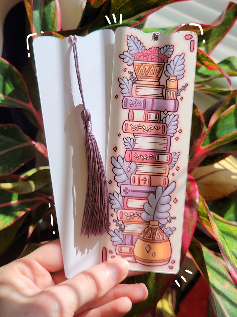 Two cute cardstock paper bookmarks with tassel and cat ear cut on the top held in a hand with a plant behind. One bookmark has an illustration of aesthetic stacked pink and purple books surrounded by pretty plants and the other the white backside.