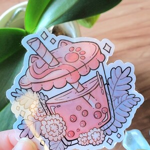 K&ouml;nnte beinhalten: Ein rosa-wei&szlig;er Sticker mit einem Boba-Tee mit einem Katzenpfoten-Deckel und einem Strohhalm. Der Boba-Tee ist von Blumen und Bl&auml;ttern umgeben.
