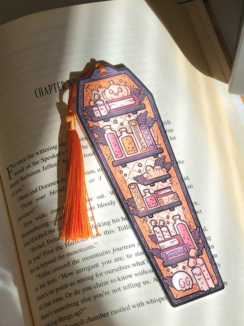 Gold orange coffin shaped bookcase bookmark with orange tassel. Features books, potions, pumpkin, candles and other witch library designs on the shelves. It rests on an open book.