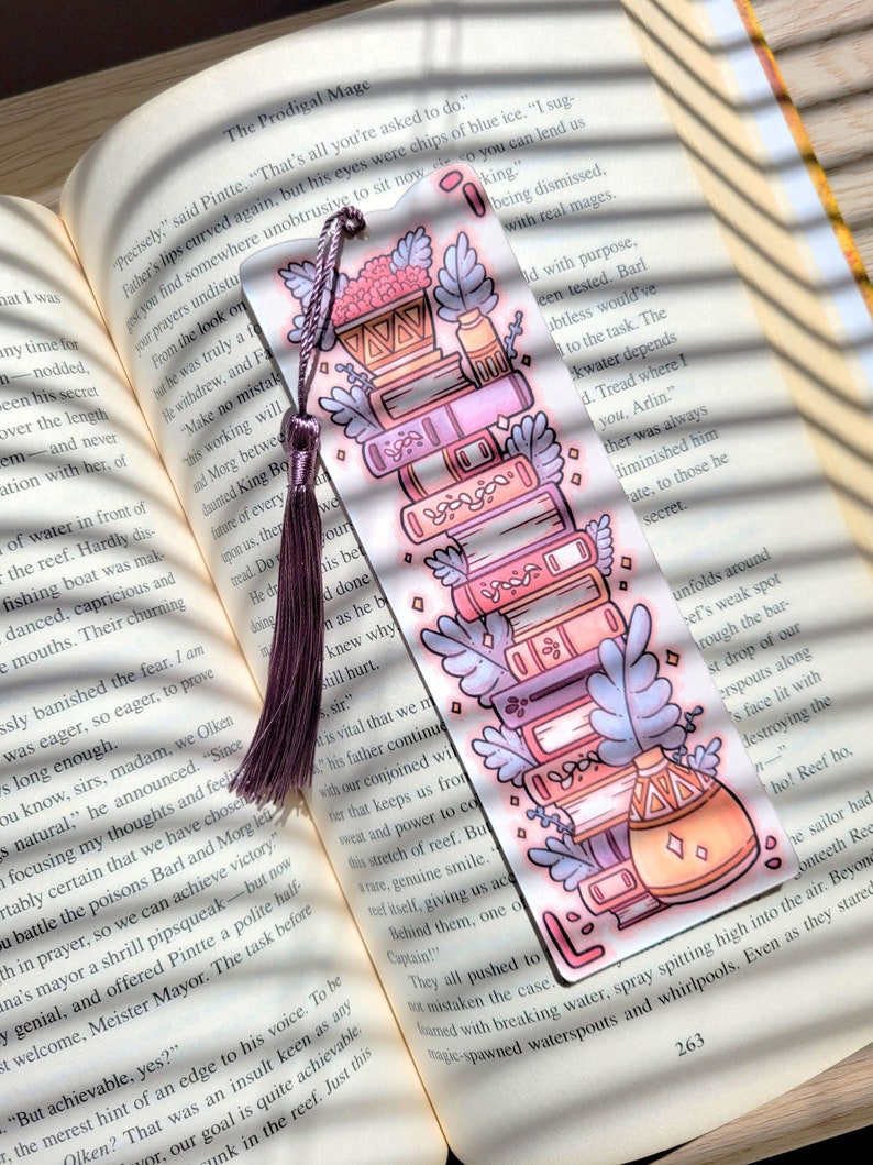 Cute cardstock paper bookmark with tassel and cat ear cut on the top resting on an open book. The bookmark has an illustration of aesthetic stacked pink and purple books surrounded by pretty plants.