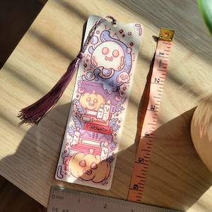Cute cardstock paper bookmark with tassel and cat ear cut on top of a light wood table. The bookmark has an illustration of stacked books, pumpkins, ghosts, and cauldron surrounded by plants. Two tape measures sit beside to show it's scale.