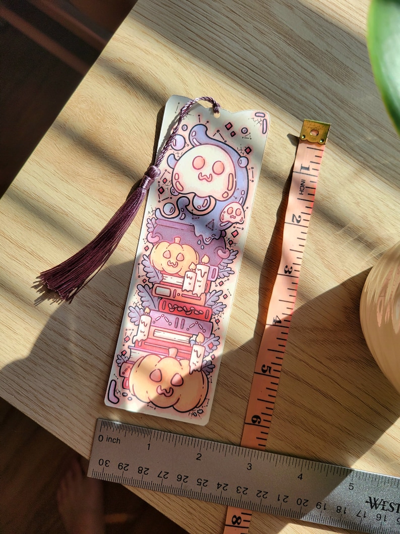 Cute cardstock paper bookmark with tassel and cat ear cut on top of a light wood table. The bookmark has an illustration of stacked books, pumpkins, ghosts, and cauldron surrounded by plants. Two tape measures sit beside to show it's scale.