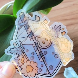 May include: A clear sticker with a cartoon illustration of a blue milk carton with a straw, flowers, and a lemon slice. The sticker has a glossy finish.