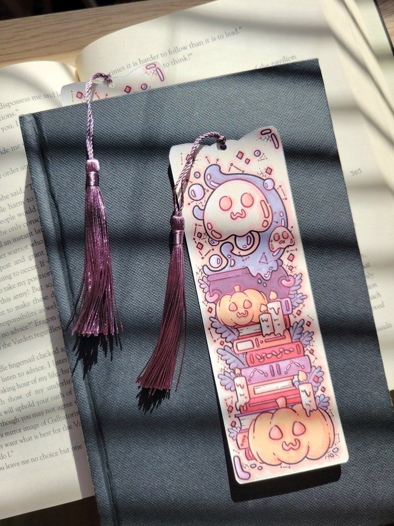 Cute cardstock paper bookmark with tassel and cat ear cut on the top resting on a black book. The bookmark has an illustration of stacked books, pumpkins, ghosts, and cauldron surrounded by plants. The black book is on an open book.