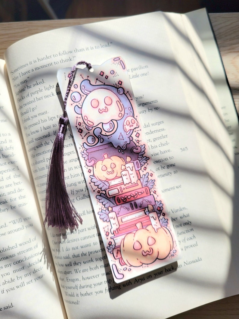 Cute cardstock paper bookmark with tassel and cat ear cut on the top resting on an open book. The bookmark has an illustration of stacked books, pumpkins, ghosts, and cauldron surrounded by plants.