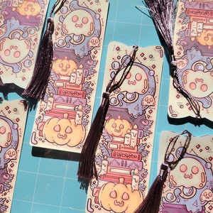 Cute cardstock paper bookmark with tassel and cat ear cut on the top resting on a purple mat. The bookmark has an illustration of stacked books, pumpkins, ghosts, and cauldron surrounded by plants. Around it are more of the same bookmark.