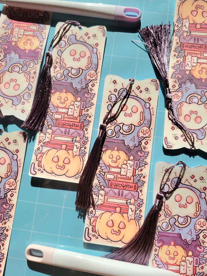 Cute cardstock paper bookmark with tassel and cat ear cut on the top resting on a purple mat. The bookmark has an illustration of stacked books, pumpkins, ghosts, and cauldron surrounded by plants. Around it are more of the same bookmark.