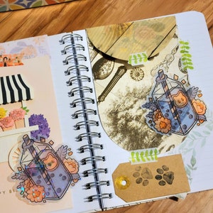 May include: A close-up of an open notebook with two stickers of a glass bottle with a straw and a cartoon bear face. The bottle is filled with a blue liquid and has a floral design. The notebook has a spiral binding and is open to a page with a brown paper tag with paw prints.
