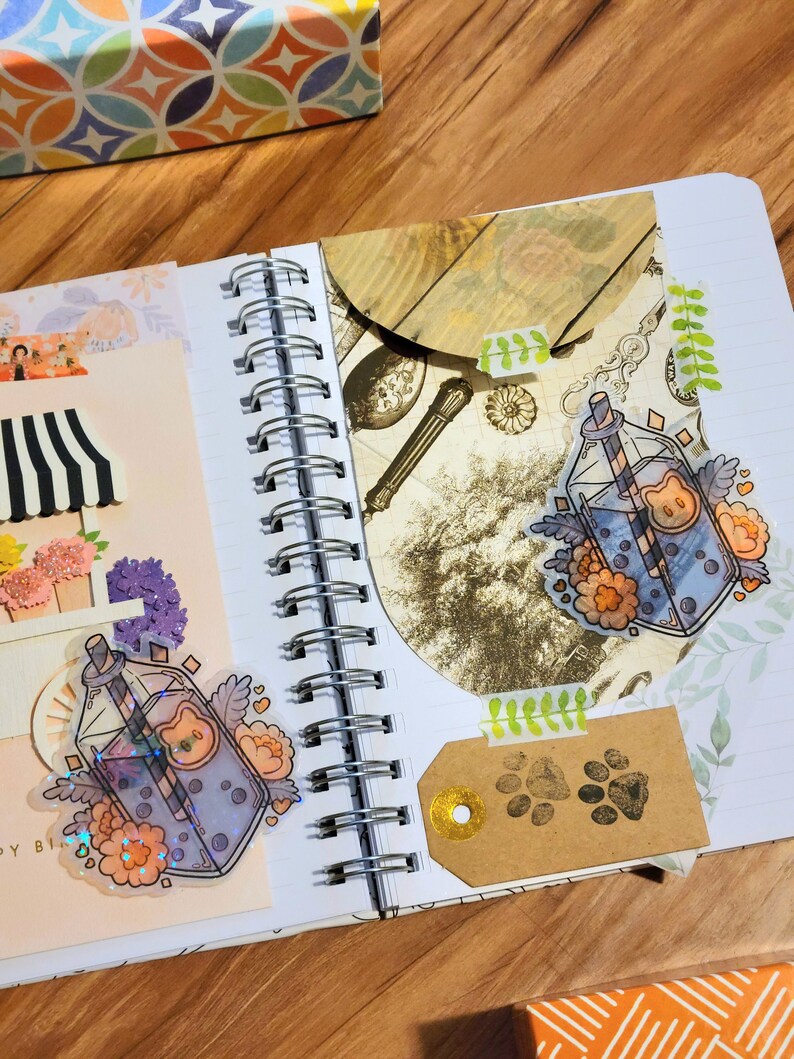 May include: A close-up of an open notebook with two stickers of a glass bottle with a straw and a cartoon bear face. The bottle is filled with a blue liquid and has a floral design. The notebook has a spiral binding and is open to a page with a brown paper tag with paw prints.