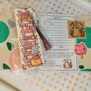 Cute cardstock paper bookmark with tassel and cat ear cut on the top resting on an packaging materials: receipt, sticker, tissue paper and card. The bookmark has an illustration of aesthetic stacked pink and purple books surrounded by pretty plants.