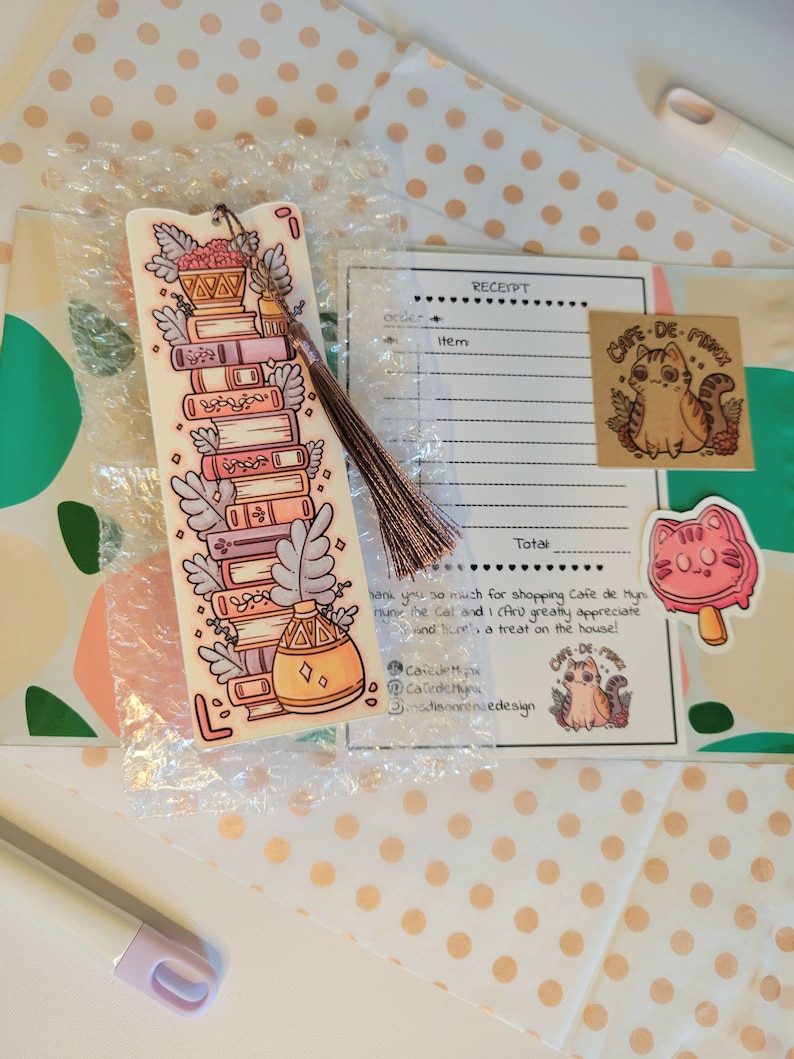 Cute cardstock paper bookmark with tassel and cat ear cut on the top resting on an packaging materials: receipt, sticker, tissue paper and card. The bookmark has an illustration of aesthetic stacked pink and purple books surrounded by pretty plants.