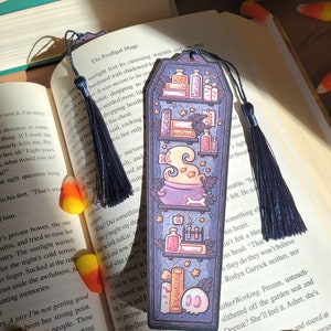 Navy blue coffin shaped bookcase bookmark with grey blue tassel. Features cauldron, skull, books, potions, and other witch library designs on the shelves. It sits on an open book with candy corn spread on the pages. Another bookmark is in the pages.