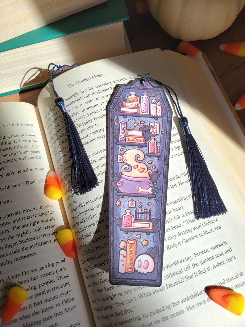 Navy blue coffin shaped bookcase bookmark with grey blue tassel. Features cauldron, skull, books, potions, and other witch library designs on the shelves. It sits on an open book with candy corn spread on the pages. Another bookmark is in the pages.