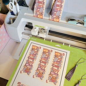 Cute cardstock paper bookmark with tassel and cat ear cut on the top resting on a green mat to be cut on a cricut maker 3 machine. The bookmark has an illustration of aesthetic stacked pink and purple books surrounded by pretty plants.