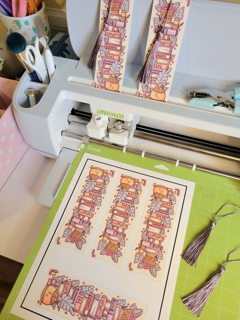 Cute cardstock paper bookmark with tassel and cat ear cut on the top resting on a green mat to be cut on a cricut maker 3 machine. The bookmark has an illustration of aesthetic stacked pink and purple books surrounded by pretty plants.