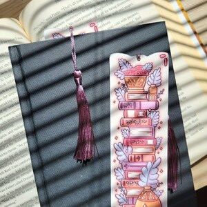 Cute cardstock paper bookmark with tassel and cat ear cut on the top resting on a black book. The bookmark has an illustration of aesthetic stacked pink and purple books surrounded by pretty plants. The black book is on an open book.