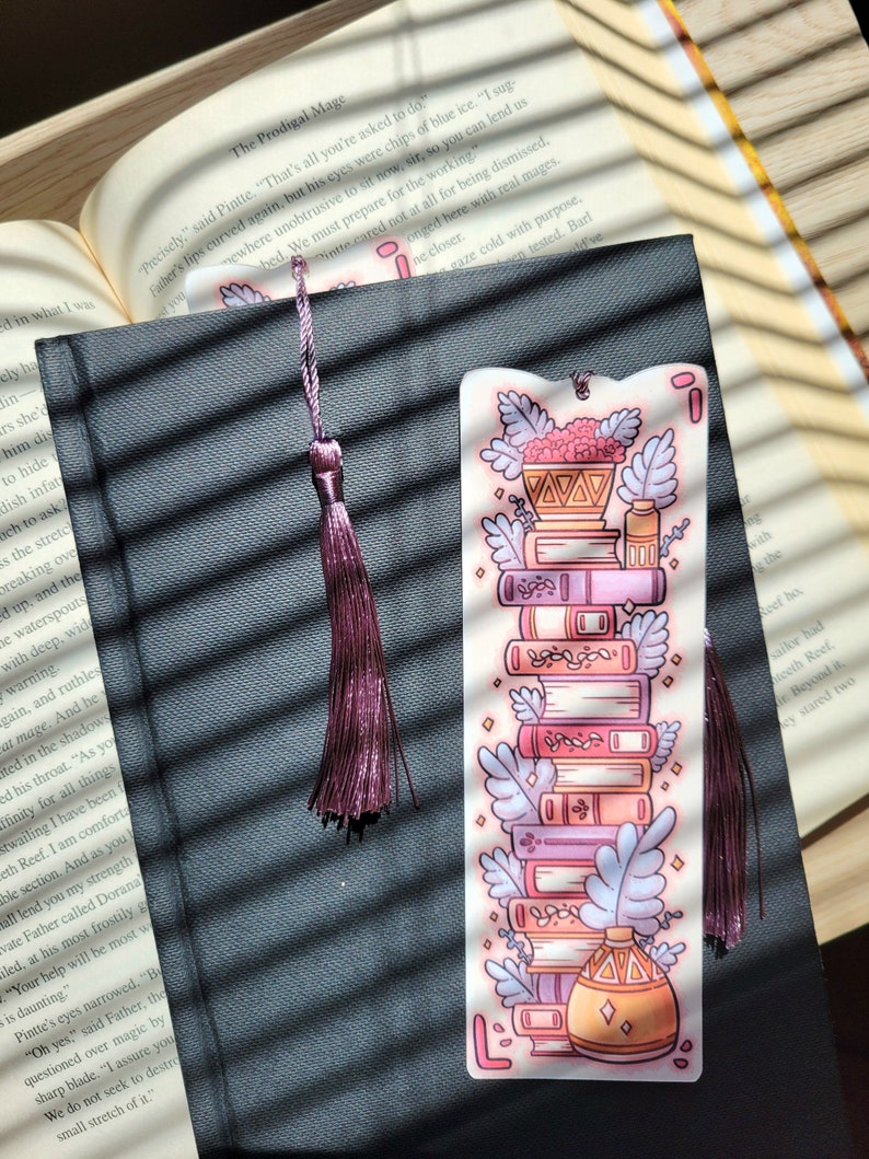 Cute cardstock paper bookmark with tassel and cat ear cut on the top resting on a black book. The bookmark has an illustration of aesthetic stacked pink and purple books surrounded by pretty plants. The black book is on an open book.