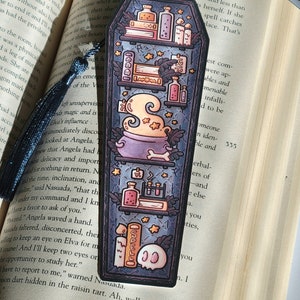 Navy blue coffin shaped bookcase bookmark with grey blue tassel. Features cauldron, skull, books, potions, and other witch library designs on the shelves. It sits on an open book.