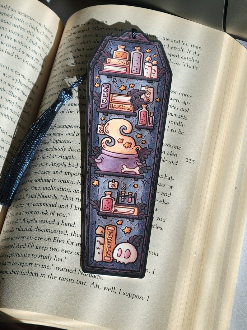 Navy blue coffin shaped bookcase bookmark with grey blue tassel. Features cauldron, skull, books, potions, and other witch library designs on the shelves. It sits on an open book.