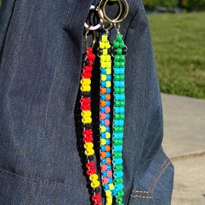 May include: Close-up of colorful beaded keychains hanging from a denim jacket. The keychains feature red, yellow, black, blue, green, and orange beads. The keychains are attached to gold and pink rings.