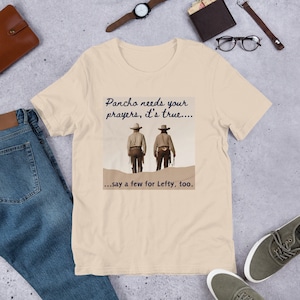 May include: A beige t-shirt featuring a graphic print of two cowboys walking in the desert. The text on the shirt reads "Pancho needs your prayers, it's true... ...say a few for Lefty, too."