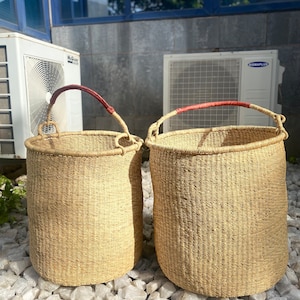 Pode incluir: Duas cestas de vime com alças de couro vermelho. As cestas são feitas de materiais naturais e têm uma cor castanho claro. Estão sentadas sobre um leito de pedras brancas.