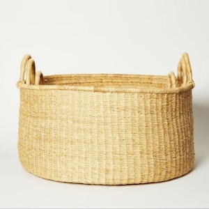 May include: A large, oval-shaped, natural woven basket with two handles on each side. The basket is made of a light brown, natural material.