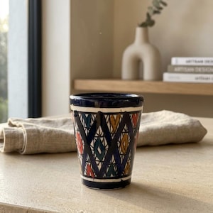 Peut inclure: Une tasse en céramique conique avec un bord et une base bleu foncé, ornée d'un motif en losanges noirs répétés. À l'intérieur des losanges se trouvent de petites formes peintes à la main en rouge, jaune et vert sur un fond crème.