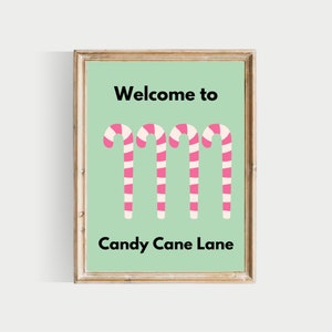 May include: A framed print with a green background and the text "Welcome to Candy Cane Lane" in black. Four pink and white candy canes are arranged vertically in the centre of the print.