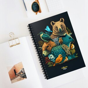 May include: A black spiral notebook with a colorful illustration of animals. The illustration features a bear, a bird, a cat, and other animals. The text "The Kingdom Says" is printed in yellow on the notebook.