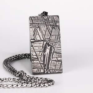 May include: A rectangular pendant with a unique geometric pattern in shades of silver and gray. The pendant is suspended from a silver chain. The design features a network of intersecting lines and shapes, creating a textured appearance. The necklace is suitable for everyday wear.
