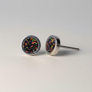 May include: A pair of stud earrings with a round, silver-colored bezel. The center of each earring features a dark background with flecks of red, green, yellow, and blue. The earrings are set against a white background.