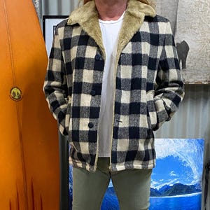 Peut inclure: Veste à carreaux noirs et beiges avec un col en fausse fourrure. La veste est boutonnée et portée sur un t-shirt blanc et un pantalon vert olive. Une planche de surf orange et un tapis coloré sont en arrière-plan.