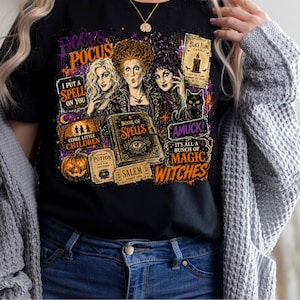 May include: A black t-shirt featuring a colorful graphic print of three women, a black cat, a pumpkin, and a book of spells. The text includes Hocus Pocus, I Put A Spell On You, Come Little Children, Black Flame Candle, Amuck, and It's All A Bunch Of Magic Witches.