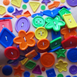 May include: A colourful bracelet made of plastic buttons in various shapes, including circles, stars, flowers, and hearts. The bracelet is worn on a person's wrist and is surrounded by loose buttons in a variety of colours.