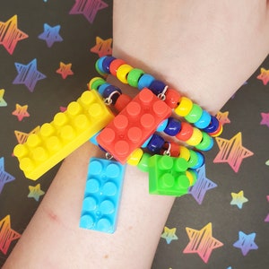 May include: A colorful beaded bracelet with four charms shaped like Lego bricks. The charms are yellow, red, blue, and green.