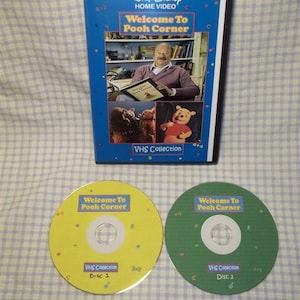 May include: A VHS collection of "Welcome to Pooh Corner" with a blue and yellow case and two yellow and green discs. The case features a photo of a man holding a book with Winnie the Pooh on the cover. The discs have the title "Welcome to Pooh Corner" and "VHS Collection" printed on them.