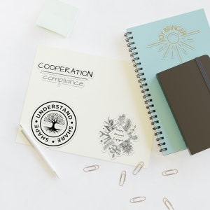 May include: An assortment of stickers and notebooks on a white surface. The stickers feature text like "COOPERATION", "UNDERSTAND", and "Happy, Relaxed, Engaged". A light blue spiral notebook and a dark brown notebook are also present, along with paper clips.