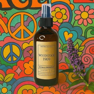 May include: A brown glass bottle of "Woodstock 1969" room and linen spray with a black cap and blue sprayer. The label is tan with black text. The background is a colorful psychedelic pattern with peace signs and flowers.