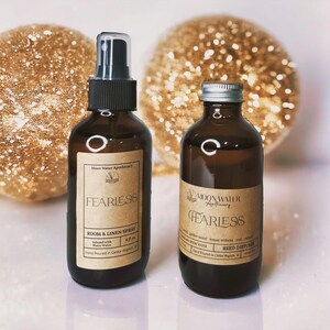 May include: Two brown glass bottles with labels that read "FEARLESS" and "Moon Water Apothecary." One is a room and linen spray, the other a reed diffuser. The background features two gold glittery spheres.