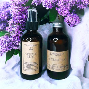 May include: Two brown glass bottles of "Vintage Lilac" room and linen spray and reed diffuser, with labels. The bottles are set against a backdrop of purple lilac flowers. The room spray bottle is 4 fl oz.