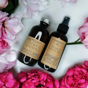 May include: Two dark brown glass bottles with labels reading "PEONY & MAGNOLIA" and "Moon Water Apothecary." One is a reed diffuser, the other a room and linen spray. The bottles are surrounded by pink peony flowers.