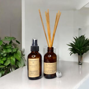 May include: Two brown glass bottles with labels that read "Rosemary & Sage" and "Room & Linen Spray". One bottle has a black spray top and the other has wooden sticks inserted into the bottle.