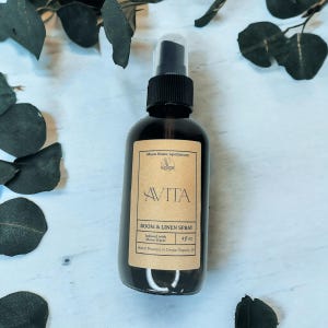 May include: A dark glass spray bottle with a black cap and a tan label. The label reads "AVITA ROOM & LINEN SPRAY" and "4 fl oz". The bottle is labeled "Moon Water Apothecary" and "Hand Poured in Cedar Rapids, IA".